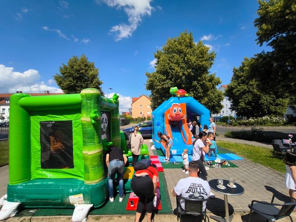 Bunte Hüpfburgen auf einem Platz mit Menschen, Bäume und blauer Himmel im Hintergrund.